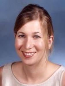A woman with light brown hair tied back smiles gently against a plain blue-gray background.