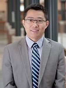 A man wearing glasses, a gray suit, white shirt, and striped tie stands indoors in a hallway with blurred columns behind him.