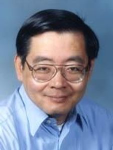 A man with short black hair and glasses smiles gently while wearing a light blue collared shirt against a plain blue.