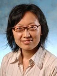 A woman with short black hair and glasses smiles slightly against a blue-gray studio background.