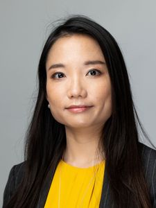 A woman with long black hair wearing a yellow top and dark pinstripe blazer looks directly at the camera against a gray.