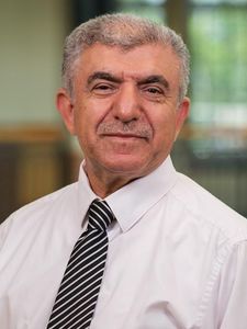 A man with gray hair and a mustache wearing a white shirt and striped tie looks directly at the camera indoors.