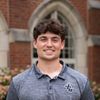 A young man with dark hair smiles at the camera wearing a gray polo shirt with a JCU logo standing outdoors.