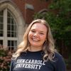 A smiling young woman with wavy blonde hair wearing a dark shirt with John Carroll University printed on it stands outside.
