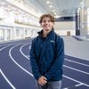 A young man wearing a navy jacket stands smiling on an indoor running track with a high ceiling and large windows behind him.