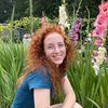 A young woman with curly red hair and glasses smiles while sitting among tall pink, white, and purple flowers outdoors.