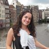 A young woman with curly hair smiles while standing on a bridge over a canal with tall, narrow buildings behind her.