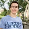 A young man wearing glasses and a John Carroll University sweatshirt smiles outdoors near a bronze statue of a kneeling.