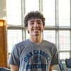 A young man with curly hair smiles standing indoors in front of large windows wearing a gray John Carroll University t-shirt.