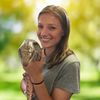 A young woman with light brown hair smiles while holding a large lizard against a blurred green outdoor background.