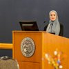 A woman wearing a gray hijab stands behind a wooden podium with a microphone and computer in front of a dark blue wall.