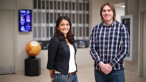 A woman in a black jacket and a man in a plaid shirt stand side by side indoors, both smiling at the camera.