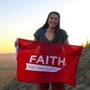 A young woman with long dark hair smiles while holding a red flag that reads "FAITH THAT DOES JUSTICE" outdoors at sunset.