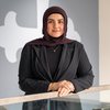 A woman wearing a dark hijab and black blazer smiles softly while leaning on a railing indoors.