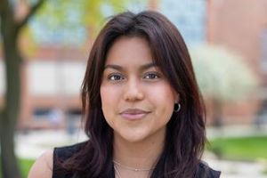 A young woman with long dark hair wearing a sleeveless black top stands outdoors with blurred trees and buildings behind her.