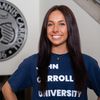 A smiling young woman with long dark hair stands with her hand on her hip wearing a John Carroll University shirt.