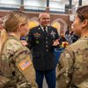 A man in a decorated military dress uniform talks and gestures with three women in camouflage uniforms inside a large hall.