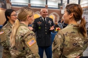 A man in a decorated military dress uniform talks and gestures with three women in camouflage uniforms inside a large hall.