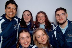 Six young adults wearing matching John Carroll University tour guide jackets smile closely together against a plain.