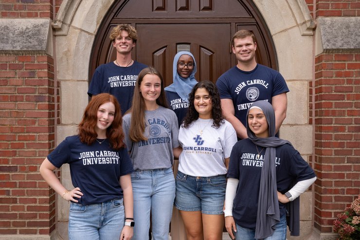 Seven diverse young adults wearing John Carroll University shirts smile and pose in front of a large wooden door.