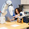 A woman with curly hair reaches toward a robotic arm on a table while working on a laptop in a lab.