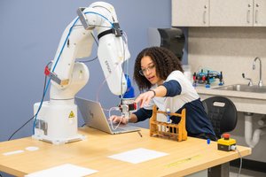 A woman with curly hair reaches toward a robotic arm on a table while working on a laptop in a lab.