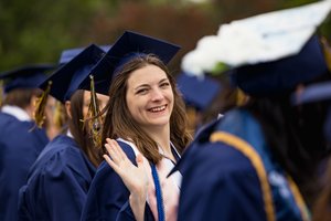 A young woman in a blue graduation cap and gown smiles and waves among a group of graduates outdoors.