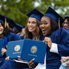 Three graduates in blue caps and gowns smile and hold up their John Carroll University diplomas outdoors.