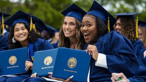 Three graduates in blue caps and gowns smile and hold up their John Carroll University diplomas outdoors.