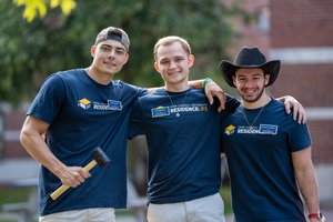 Three young men wearing matching blue John Carroll University Residence Life shirts smile with their arms around each other.