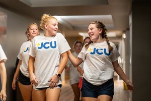 Two young women wearing white JCU t-shirts walk and smile at each other in a hallway with others behind them.