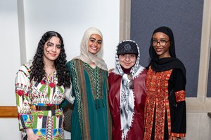 Four women stand closely together smiling, wearing colorful traditional dresses and headscarves in an indoor setting.