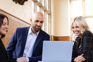 Three people in business attire smile and look at a laptop in a room with tall windows and wood paneling.