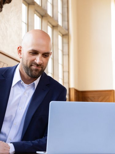 Three people in business attire smile and look at a laptop in a room with tall windows and wood paneling.