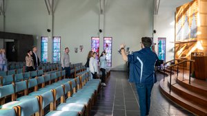 A man in a blue jacket gestures with raised hands toward a small group standing near stained glass windows in a church.