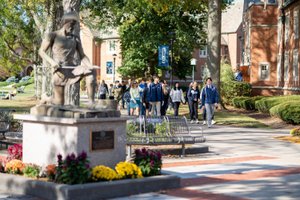 A group of people walk along a tree-lined campus path past a statue of a kneeling figure holding a sword.