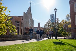 A group of people walk along a sunny campus path past a building with a large circular window and a cross on top.