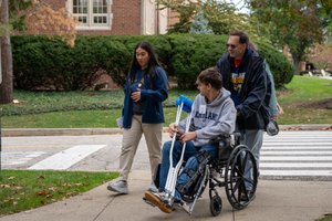 A young man with a leg brace sits in a wheelchair holding crutches while two people walk beside and behind him on a sidewalk.