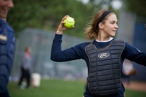 A young softball player wearing a chest protector prepares to throw a bright yellow softball during practice.
