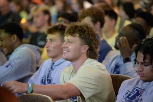 A group of young men sit closely together in an audience, with one smiling and reaching forward.