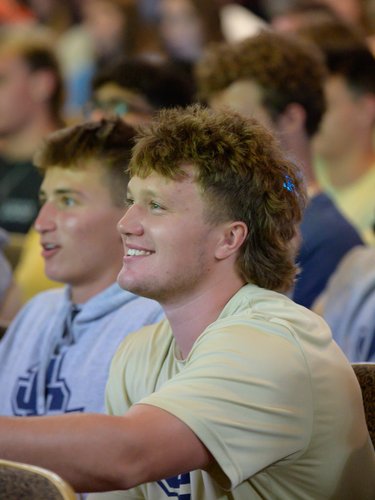 A group of young men sit closely together in an audience, with one smiling and reaching forward.