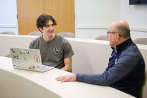 A young man with a laptop covered in stickers smiles while talking to an older man in a classroom.