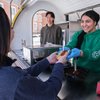 A person wearing blue gloves and a green hoodie smiles while handing a food tray to another person at a food stand.