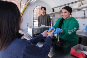 A person wearing blue gloves and a green hoodie smiles while handing a food tray to another person at a food stand.
