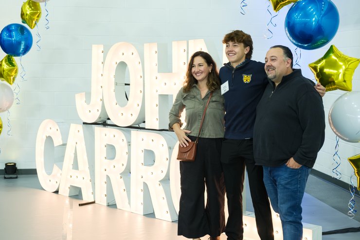 Three people smile and pose in front of large illuminated letters spelling JOHA CARRILLO with blue and gold star balloons.