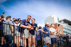 A group of young people stand behind a black railing in a stadium, some smiling and clapping under a blue sky.