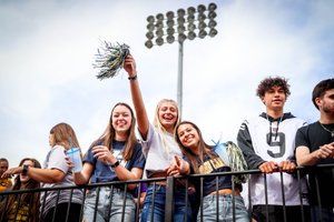 A group of young people stand behind a railing outdoors, smiling, holding drinks and pom-poms under stadium lights.