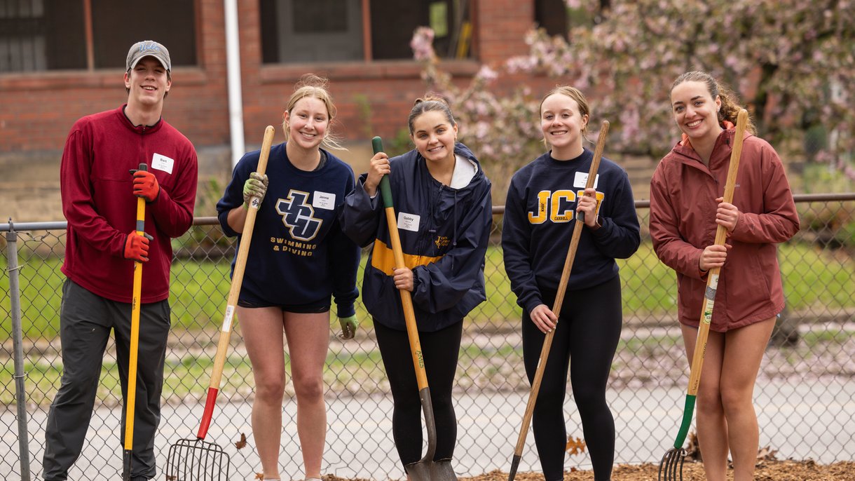 Five young people stand outside holding gardening tools and smiling in front of a chain-link fence and a brick building.