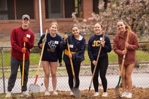 Five young people stand outside holding gardening tools and smiling in front of a chain-link fence and a brick building.