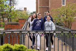 Four young women walk together on a paved path between brick buildings, smiling and wearing casual sweatshirts.